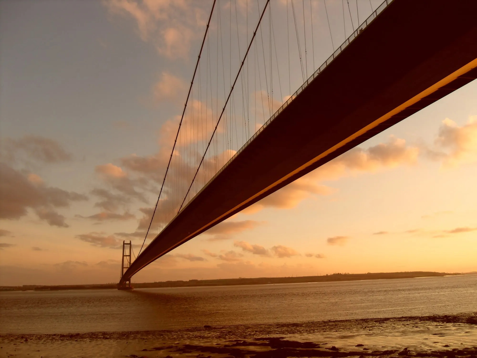 The Humber Bridge at sunset, seen from beneath the deck on the Hessle foreshore, with the suspension cables sweeping away towards the south tower over the estuary