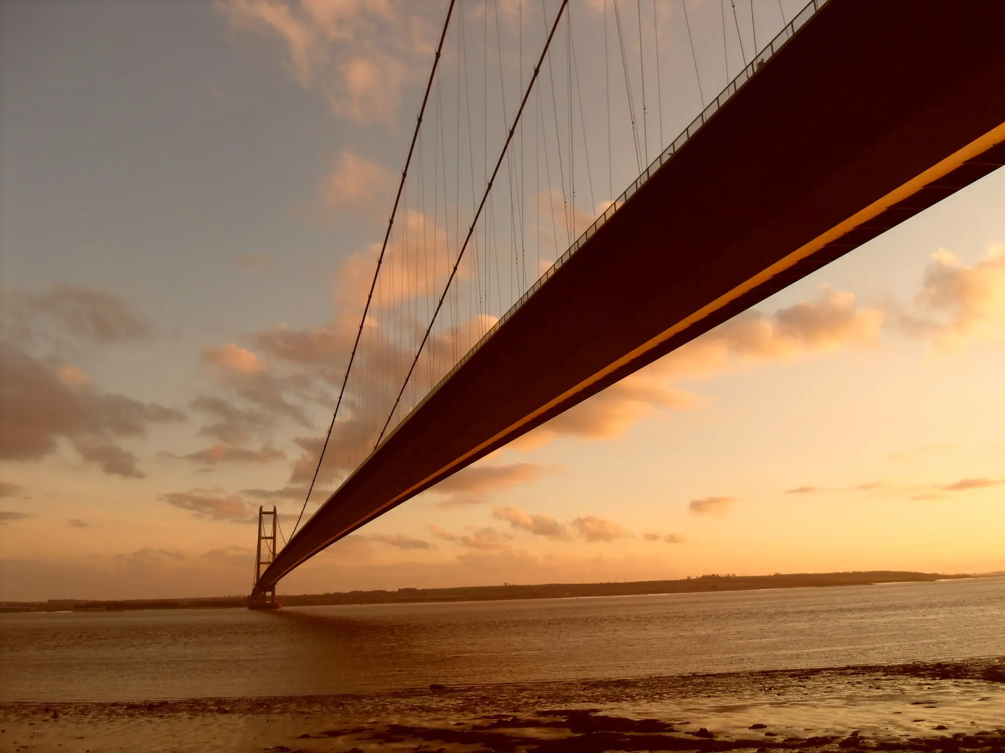 The Humber Bridge at sunset, seen from beneath the deck on the Hessle foreshore, with the suspension cables sweeping away towards the south tower over the estuary